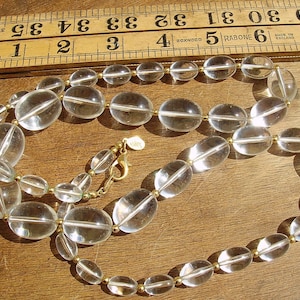 May include: A clear crystal necklace with gold-toned beads and a clasp. The necklace is made of oval-shaped beads and is laid out on a wooden surface.