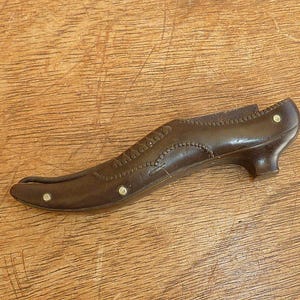 Puede incluir: Objeto marrón con forma de zapato de estilo vintage, con un tacón pequeño y costuras decorativas. El zapato tiene una punta afilada y está adornado con pequeños tachuelas redondas plateadas. El objeto está sobre un fondo de madera.