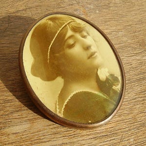 May include: Oval sepia-toned portrait of a woman with a pearl headband and dark clothing. The portrait is framed in a bronze-colored metal. The image is set against a wooden surface.