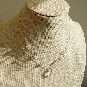 Puede incluir: Un delicado collar plateado con cuentas de cristal rectangulares transparentes. El collar se exhibe en un busto de joyería beige. Los cristales están espaciados uniformemente a lo largo de la cadena, con un grupo de cristales en el colgante.
