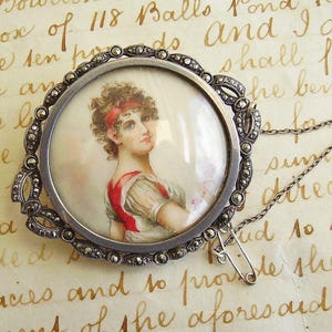May include: Antique silver-tone brooch featuring a miniature portrait of a woman with a red headband and dress accents. The brooch has a decorative marcasite-studded frame and a safety pin closure with a chain.