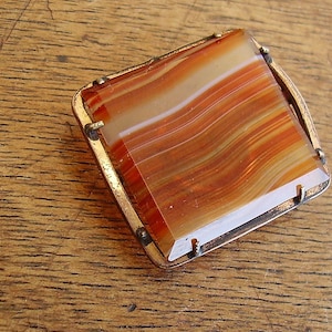 May include: A vintage agate brooch with a copper-toned metal frame. The agate is a reddish-brown colour with white stripes.
