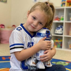 Op de afbeelding: Een jong meisje omhelst een wit en blauw R2-D2 speelgoedrobot. De robot heeft een zilveren kop en is een populair personage uit de Star Wars-franchise. Het meisje draagt een wit shirt met blauwe accenten en de letter "R".