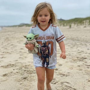 Op de afbeelding: Een kind draagt een lichtblauw shirt met de tekst "STAR MANDOALRIAN" en een afbeelding van een personage. Het kind houdt een kleine groene pop van een personage uit dezelfde franchise vast. Het shirt heeft bruine en witte strepen.
