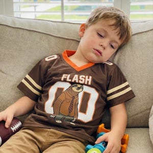 Op de afbeelding: Bruin shirt met oranje rand en witte strepen op de mouwen. Het shirt heeft het woord "FLASH" en het nummer "80" met een cartoon luiaard. Het shirt wordt gecombineerd met een kaki broek.