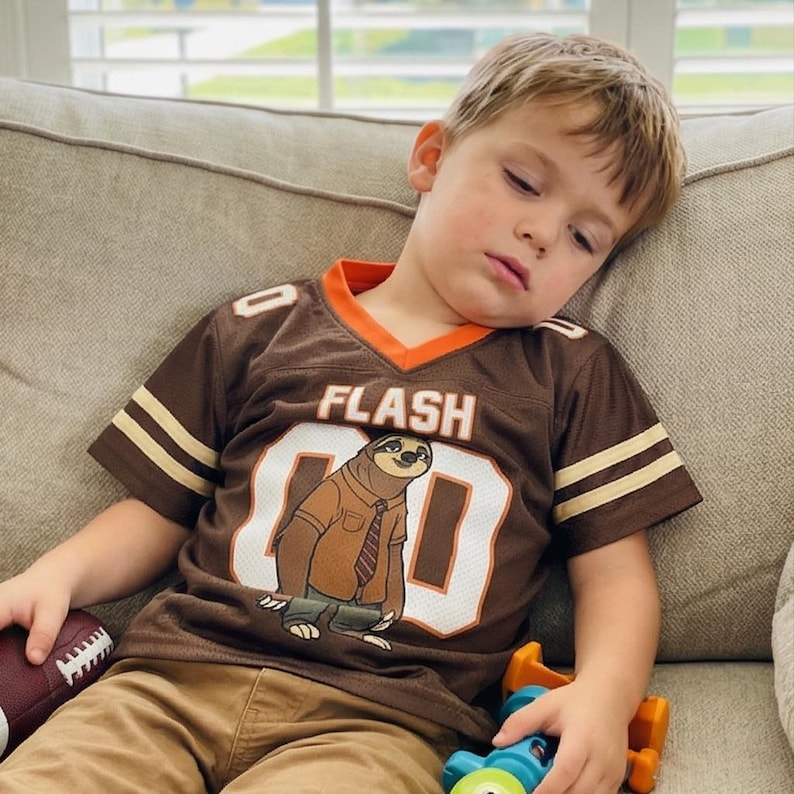 Op de afbeelding: Bruin shirt met oranje rand en witte strepen op de mouwen. Het shirt heeft het woord "FLASH" en het nummer "80" met een cartoon luiaard. Het shirt wordt gecombineerd met een kaki broek.
