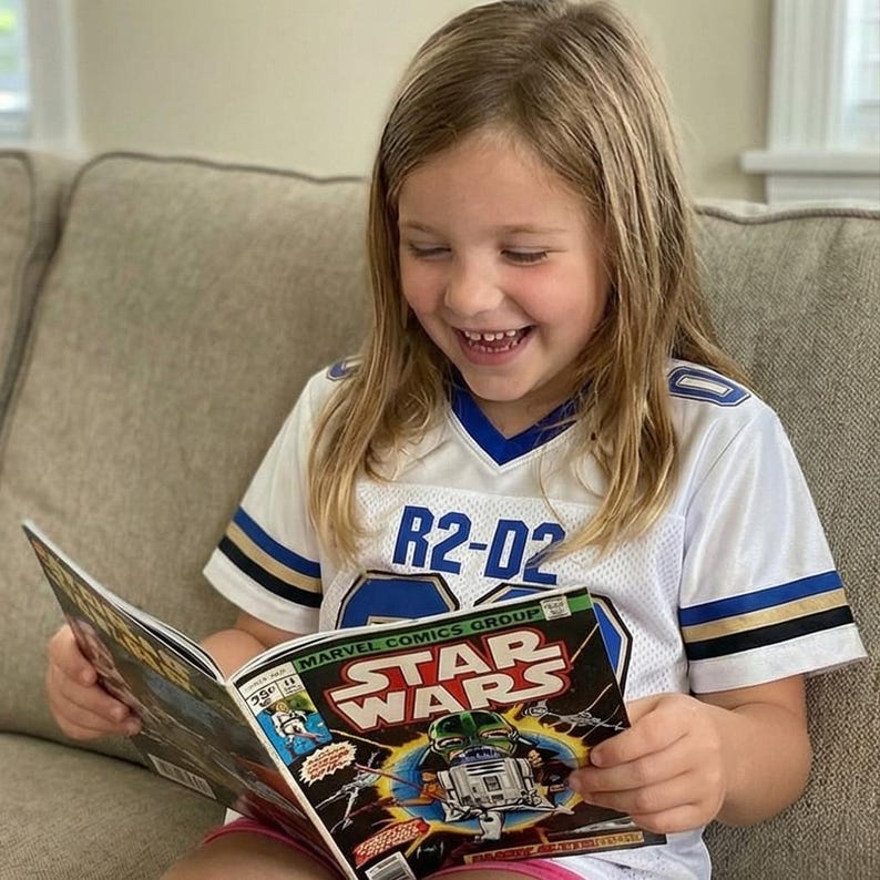Op de afbeelding: Een jong persoon draagt een wit en blauw shirt met "R2-D2" erop gedrukt, glimlacht terwijl hij een "Star Wars" stripverhaal leest. De cover van het stripverhaal toont R2-D2 en andere personages uit de Star Wars-franchise.