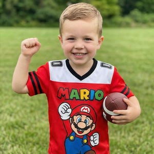 Pu&ograve; includere: Un bambino indossa una maglia rossa a tema Mario con dettagli bianchi e neri, tenendo in mano un pallone da football americano marrone. La maglia mostra la parola "MARIO" in verde e una grafica di Mario. Il bambino sorride e ha il pugno alzato.