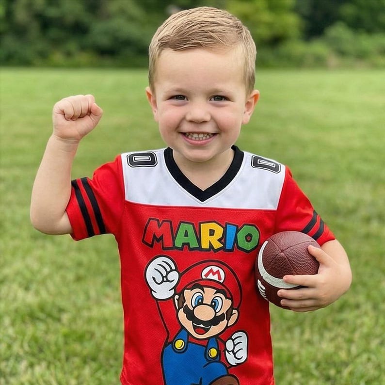Pu&ograve; includere: Un bambino indossa una maglia rossa a tema Mario con dettagli bianchi e neri, tenendo in mano un pallone da football americano marrone. La maglia mostra la parola "MARIO" in verde e una grafica di Mario. Il bambino sorride e ha il pugno alzato.