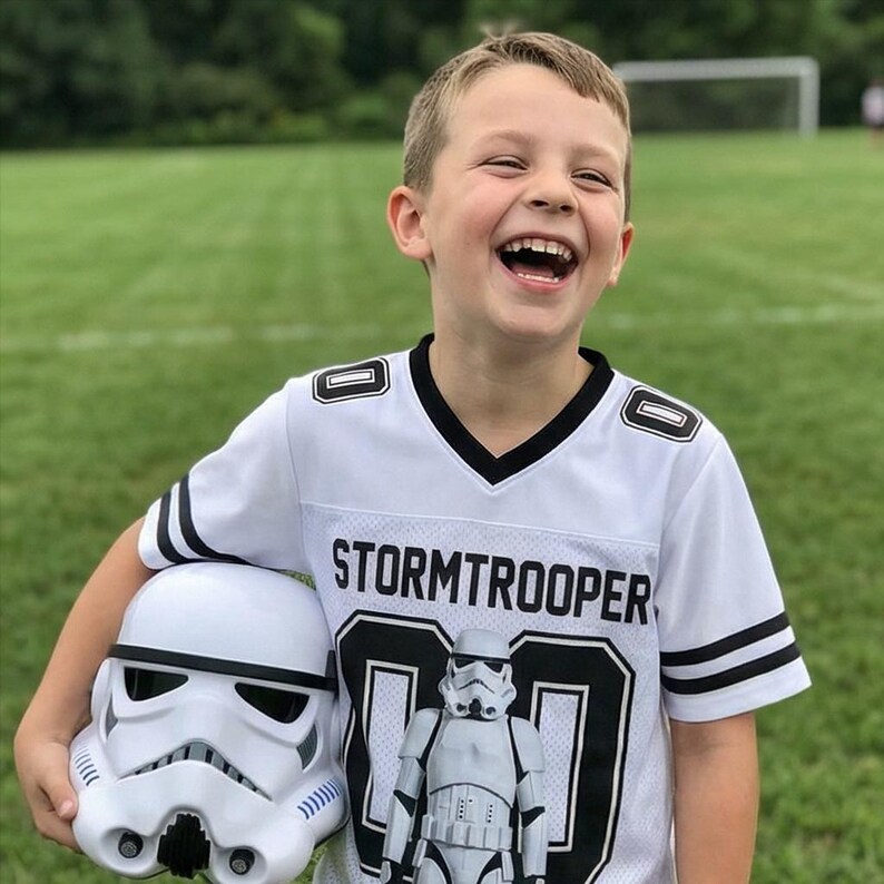Pu&ograve; includere: Una maglia bianca da Stormtrooper con dettagli neri e la scritta "Stormtrooper" stampata sul petto. La maglia ha il numero 00. Un casco bianco da Stormtrooper &egrave; tenuto tra le braccia della persona.