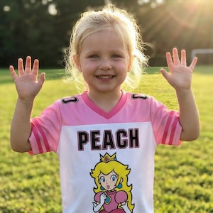 Pu&ograve; includere: Una bambina indossa una maglia rosa e bianca con la scritta "PEACH" stampata sul davanti. La maglia presenta una grafica della Principessa Peach della serie di videogiochi Super Mario. La bambina sorride e saluta la fotocamera.