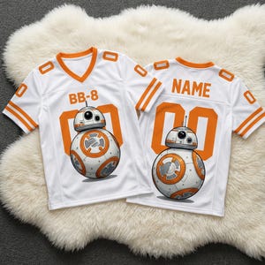 Op de afbeelding: Twee witte American football-shirts met oranje details en de afbeelding van BB-8 uit Star Wars. E&eacute;n shirt heeft "BB-8" en de andere "NAME" op de rug. De shirts liggen op een witte, zachte ondergrond.