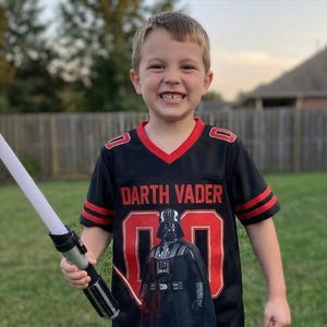 Op de afbeelding: Een kind draagt een zwart en rood Darth Vader-shirt met nummer 00. Het shirt heeft de woorden "Darth Vader" in rode letters. Het kind houdt een wit en zwart lichtzwaard vast en glimlacht.