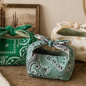 May include: Three fabric knot bags featuring traditional bandana paisley patterns in green, light teal, and cream with gold accents. Each bag has a knotted handle and a green and white checkered interior lining, constructed from soft textile material.
