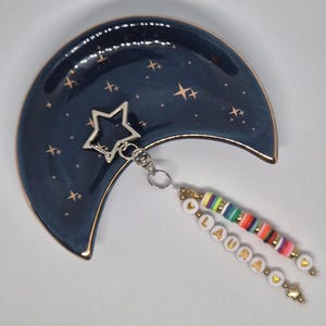 May include: A dark blue crescent moon-shaped ceramic dish with gold star accents and a metallic rim. Attached is a silver star-shaped keychain featuring two beaded strands, one with multi-colored clay discs and one spelling the name LAURA.