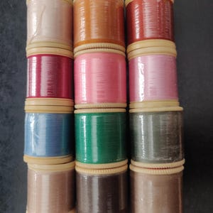 May include: A collection of twelve cylindrical spools of sewing thread arranged in a grid of three columns and four rows. The spools feature various solid colours including light pink, orange, red, magenta, bright pink, blue, green, grey, and brown.