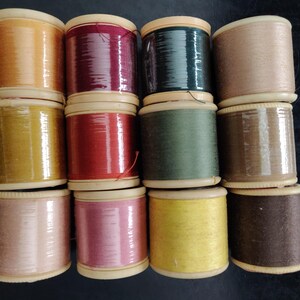 May include: A selection of vintage thread spools in a variety of colours, such as yellow, red, green, and brown. The spools are made of wood and wrapped with thread, creating a colourful display. The spools are arranged in a grid pattern.