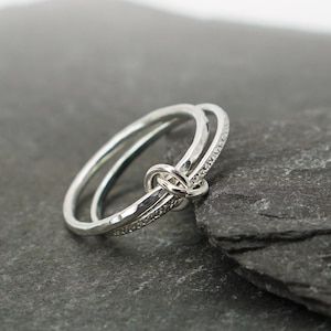 May include: Two silver rings, one with a textured finish and the other with a smooth finish, are intertwined and knotted together. The rings are displayed on a dark grey stone surface.