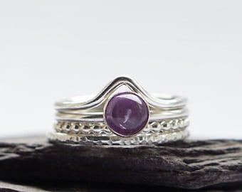 May include: A set of four silver rings with a purple gemstone in the center. The rings are stacked on top of each other and are on a gray stone surface.