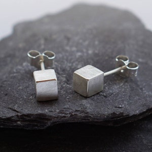 May include: A pair of silver cube stud earrings on a dark grey stone surface.