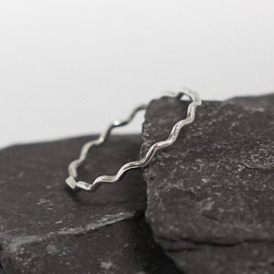 May include: A silver ring with a wavy, textured design. The ring is resting on a dark grey stone.