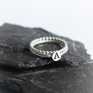 May include: A silver ring with a beaded design and a small charm with the letter 'A' on it. The ring is resting on a piece of black slate.
