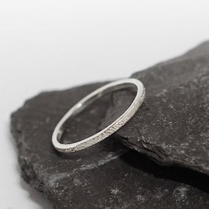 May include: A thin silver band ring with a textured surface, resting on two pieces of dark gray slate.