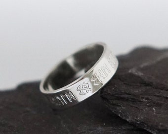 May include: A silver ring with a cross design and an inscription around the band. The ring is sitting on two pieces of dark gray slate.