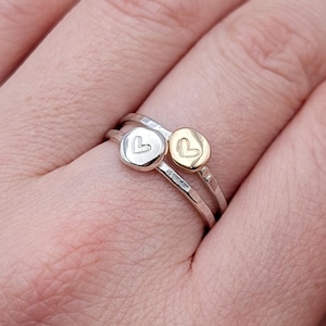 May include: Two silver rings with hammered bands, one with a silver heart-shaped charm and the other with a gold heart charm. The rings are stacked on a person's finger, showcasing the jewelry's design and detail.