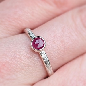 May include: A delicate silver ring featuring a small, faceted ruby gemstone set in the centre. The band is thin and has a textured finish.