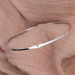 May include: A silver bangle bracelet with a hammered finish. The bracelet has a single initial "F" engraved on the front.