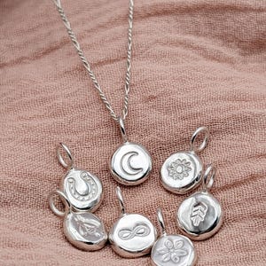 May include: A silver necklace with a fine chain and a crescent moon pendant. Several silver charms with designs like a horseshoe, sun, leaf, flower, and infinity symbol are scattered around. The jewellery is displayed on a pink fabric background.
