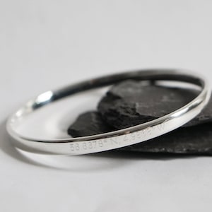 May include: Silver bangle bracelet with a smooth, rounded surface. The bracelet is engraved with the coordinates "58.8878° N, 2.9848° W".