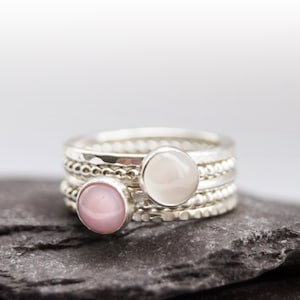 May include: A set of four silver stacking rings with pink and white gemstones. The rings are on a grey stone surface.