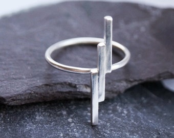 May include: A silver ring with three vertical bars extending upwards from the band. The ring is resting on a dark gray stone.