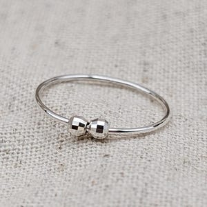 May include: A delicate silver ring featuring two small, textured beads. The ring has a thin band and a minimalist design. The jewelry is displayed on a neutral, textured surface, highlighting its simple elegance.
