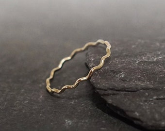 Pode incluir: Um anel fino de cor dourada com um design de borda ondulada ou festonada contínua. A banda é delicada e circular, apresentando um padrão curvo repetitivo ao longo de toda a sua circunferência, criando uma textura ondulada.