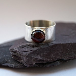 May include: A silver ring with a round, dark red gemstone set in the centre. The ring is thick and has a hammered texture.