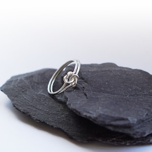 May include: A silver-colored ring featuring a delicate knot design at the center. The band is constructed from two thin, parallel wires that converge into a central knot detail, creating a continuous and smooth metallic loop.