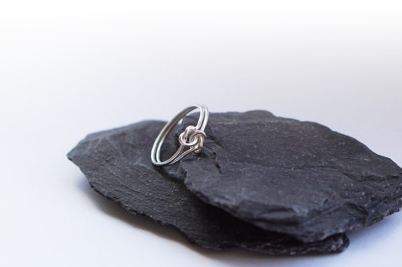 May include: A silver-colored ring featuring a delicate knot design at the center. The band is constructed from two thin, parallel wires that converge into a central knot detail, creating a continuous and smooth metallic loop.