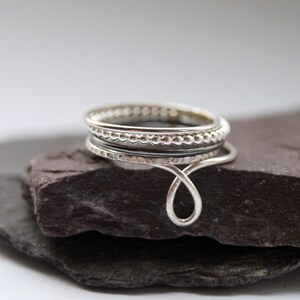 May include: A silver ring with a unique design featuring multiple bands and a curved wire detail. The ring is displayed on a dark grey stone.