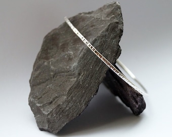 May include: A silver bangle bracelet with a textured surface. The bracelet is displayed on a dark gray rock.