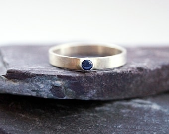 Puede incluir: Un anillo plateado con una pequeña gema redonda de color azul oscuro engastada en la banda. El anillo se muestra sobre una superficie de piedra gris oscuro y texturizada sobre un fondo blanco. La banda del anillo es lisa y plana.