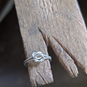 May include: A silver-colored metal ring featuring a central knot design made from a single continuous wire. The ring is positioned around the end of a textured, light brown wooden bench pin with visible grain and cracks.