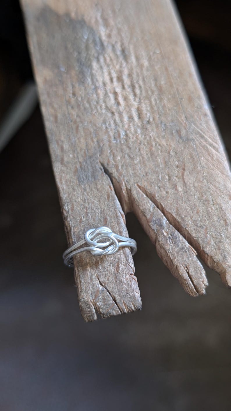 May include: A silver-colored metal ring featuring a central knot design made from a single continuous wire. The ring is positioned around the end of a textured, light brown wooden bench pin with visible grain and cracks.