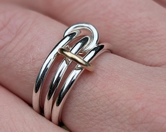 May include: A multi-band ring featuring three polished silver bands that are interconnected and secured by a single, smooth gold-toned horizontal bar. The design creates a layered, looped appearance with a reflective metallic finish on all components.