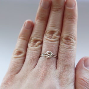 May include: A silver-colored ring featuring a delicate wire-wrapped knot design at the center. The band is constructed from a thin, smooth metallic wire that loops into a central knot, creating a continuous and minimalist circular shape.