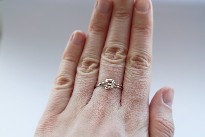 May include: A silver-colored ring featuring a delicate wire-wrapped knot design at the center. The band is constructed from a thin, smooth metallic wire that loops into a central knot, creating a continuous and minimalist circular shape.