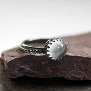 May include: A silver ring featuring a white gemstone set in an intricate, ornate design. The ring is displayed on a dark grey stone surface.