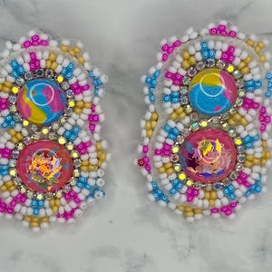 Puede incluir: Par de pendientes de cuentas con un diseño colorido. Cada pendiente presenta una piedra redonda superior con un remolino de azul, amarillo y rosa, y una piedra rosa inferior con purpurina iridiscente. Los pendientes están rodeados de cuentas blancas, amarillas, azules y rosas.
